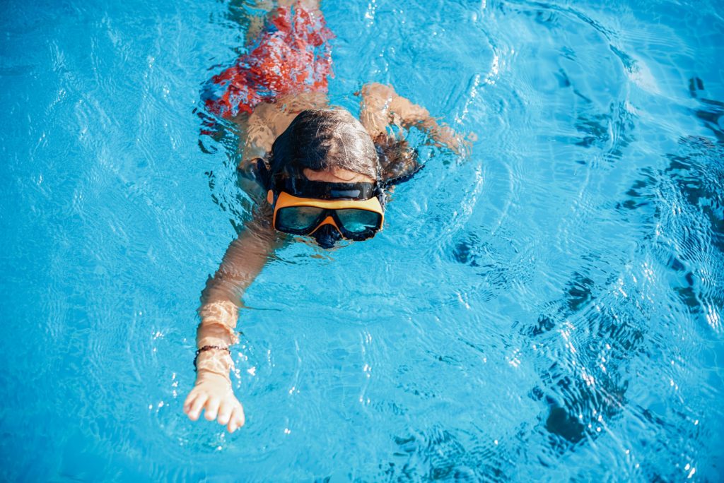 Sportive kid with goggles swims in pool and pose on camera